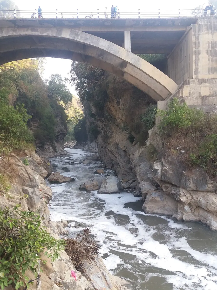 Looking along a narrow gorge with steep, gray rock walls and trees on top, with a fast, foaming river at the bottom, under an arched concrete bridge