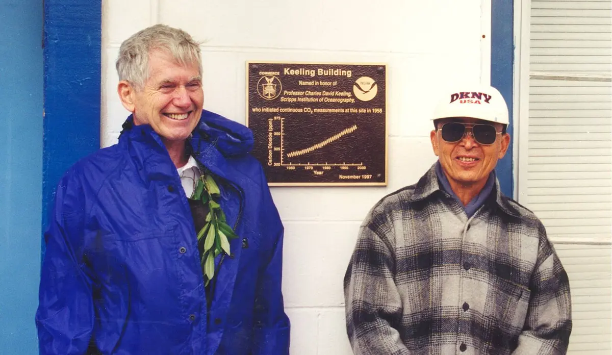 Two smiling men standing by a white wall, on either side of a bronze plaque headed Keeling Building, with a small graph of a zigzag line climbing from 1960 to 2000