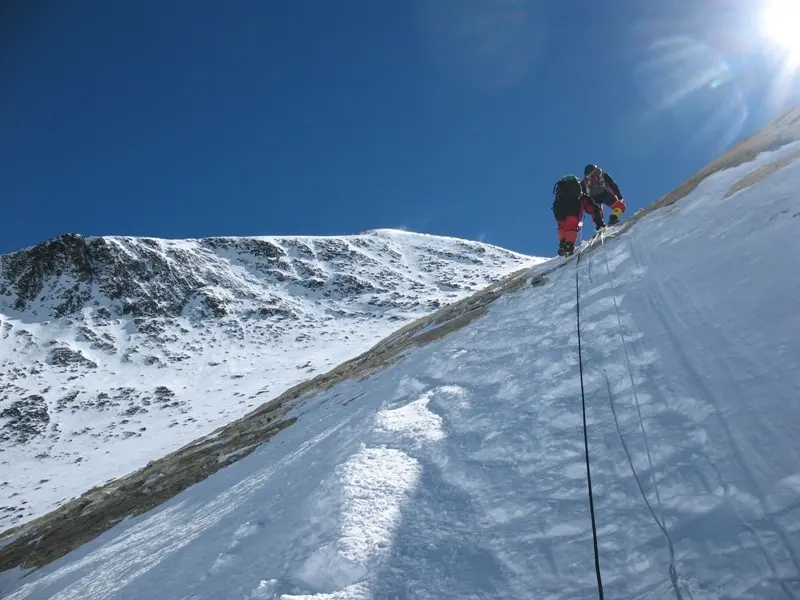 A climber in a red suit on a steep snow slope, clipped to a rope, crossing a band of brownish-yellow rock that runs diagonally across the snow under a deep blue sky