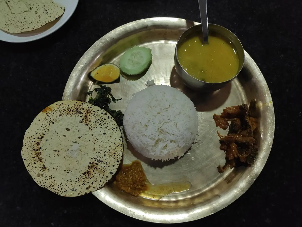 A brass plate seen from above, with a mound of white rice in the middle, a bowl of yellow dal, a papad, fried meat, pickle, cucumber, lime and a little saag