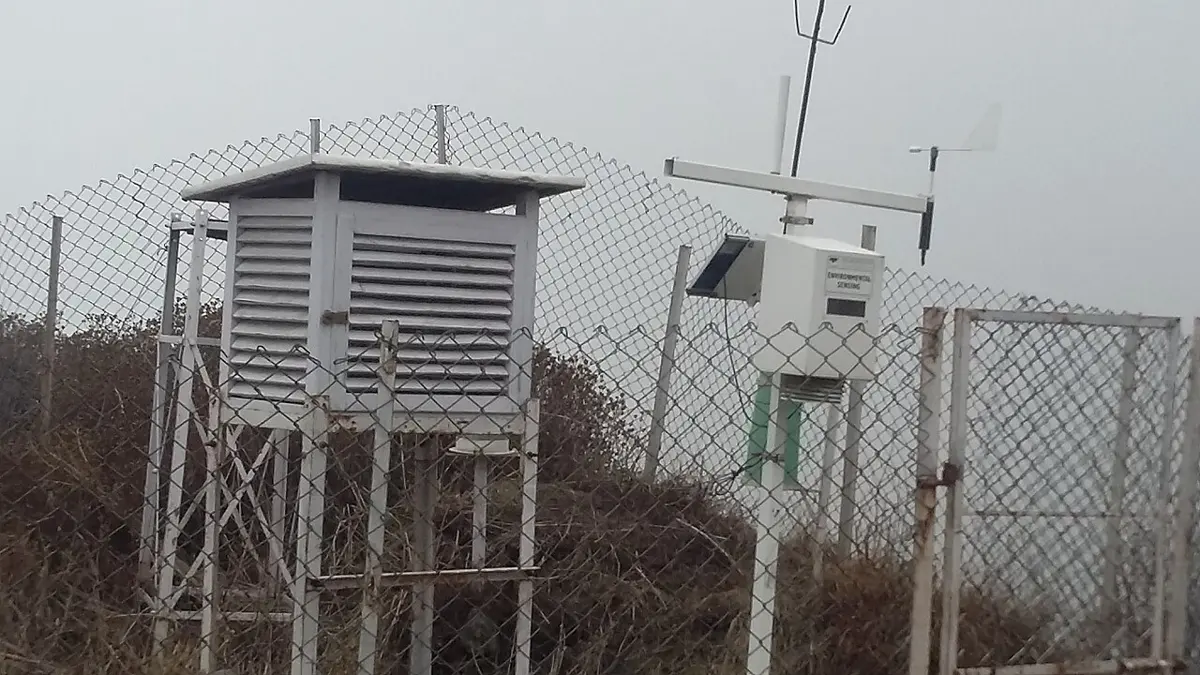 Behind a wire fence on a misty hillside, a white wooden box with slatted sides on legs, beside a white automatic weather station on a pole with a solar panel and a wind vane with a small propeller on its nose