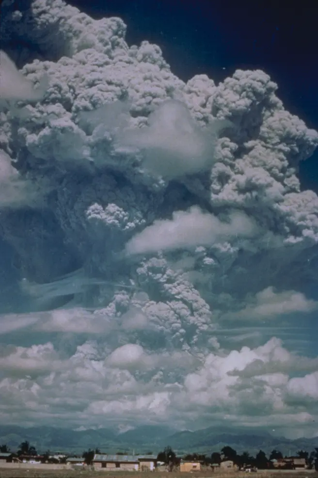 A towering gray column of ash billowing up above white clouds, with a row of low buildings and trees far below it