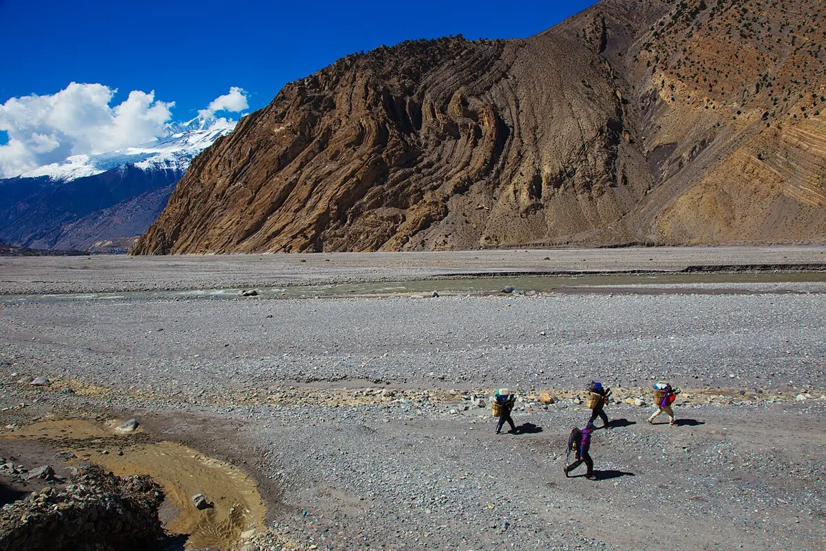 Porters with baskets walking along the wide, stony bed of the Kali Gandaki; behind them a bare brown mountainside shows rock layers bent into tight curves