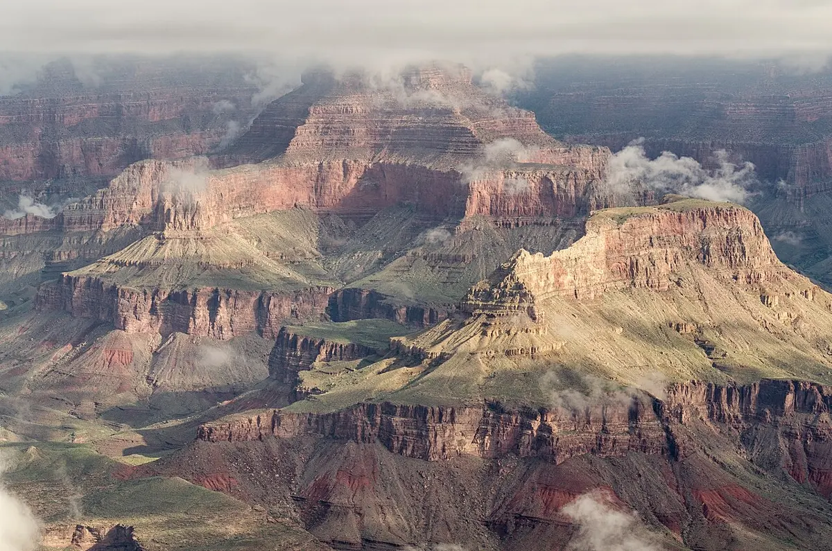 A huge canyon with steep walls made of many horizontal bands of red, brown and cream rock, with green slopes between the cliffs