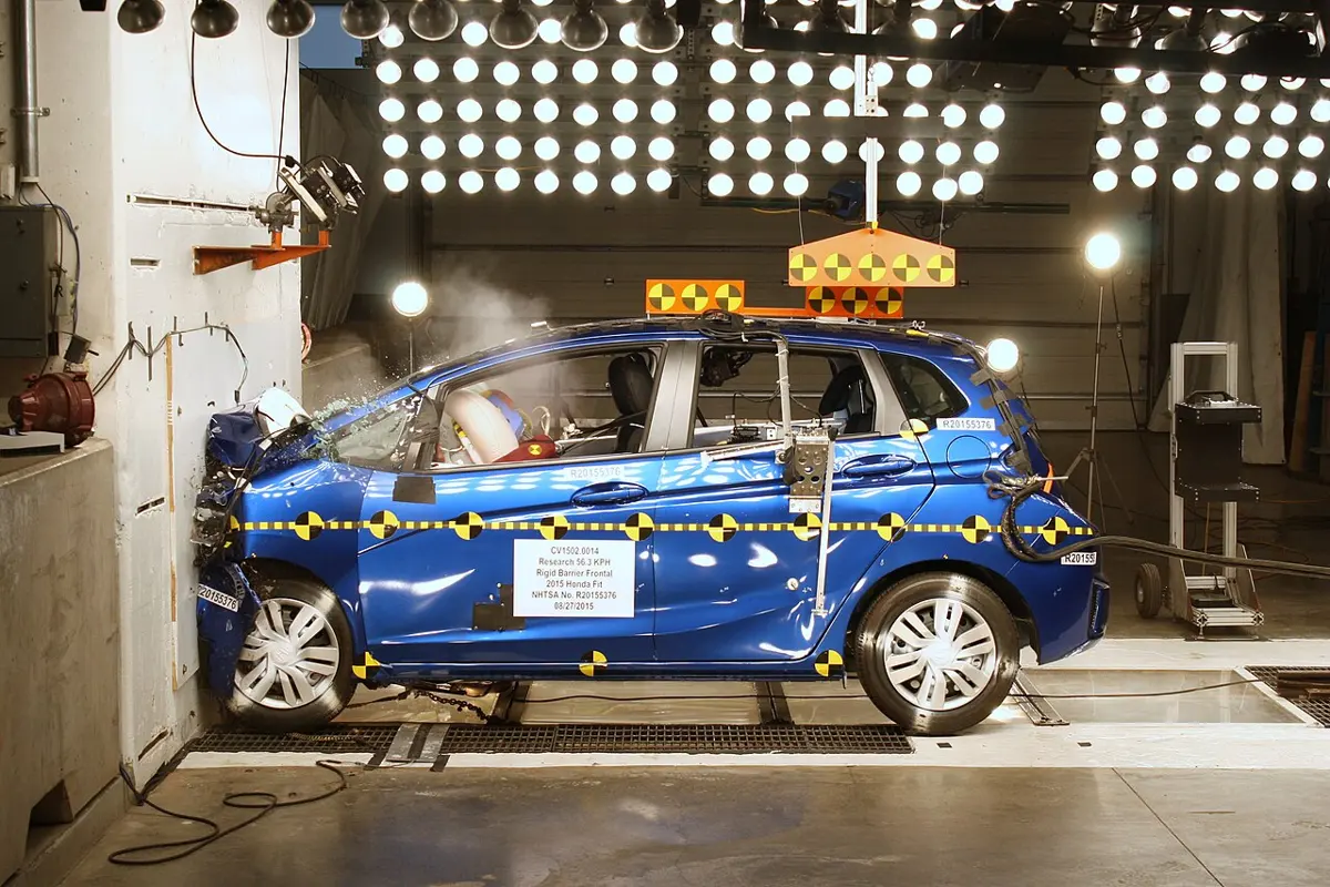 A small blue car pressed against a concrete wall in a brightly lit test hall; its front is crumpled and its airbag has inflated, while the doors and roof are undamaged