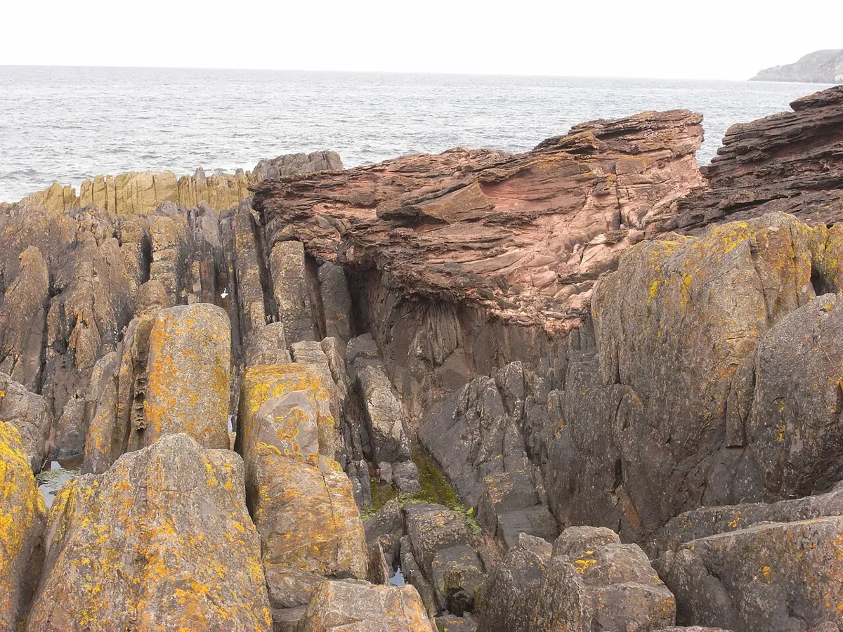 A rocky shore by the sea: gray layers of rock stand almost upright in the front, and above them, in the middle, sloping layers of red sandstone lie across their tops