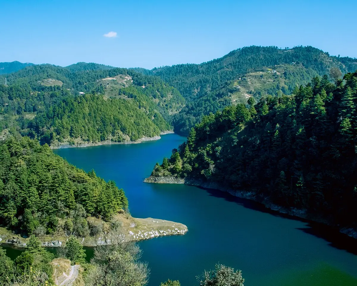 A long, deep blue lake winding between steep hills covered in pine forest, under a clear sky