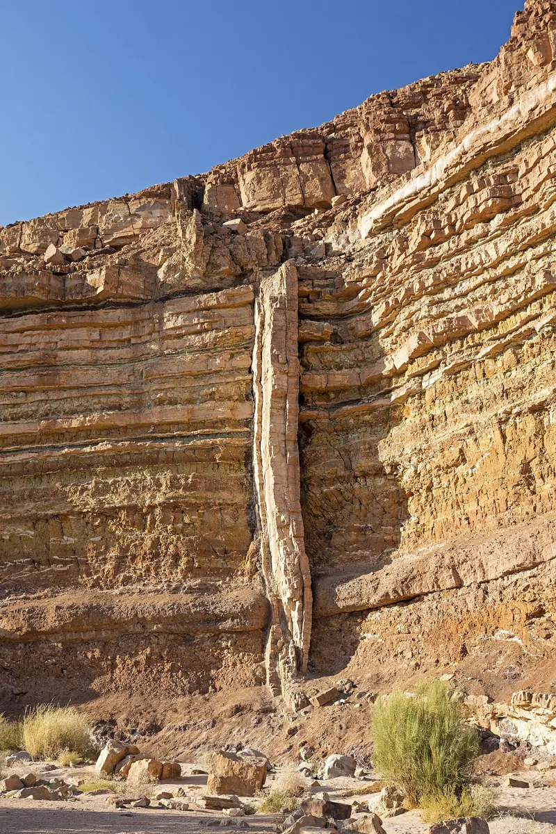 A desert cliff of flat, buff and brown rock layers, cut by a pale, upright band of rock that runs straight up through them like a wall