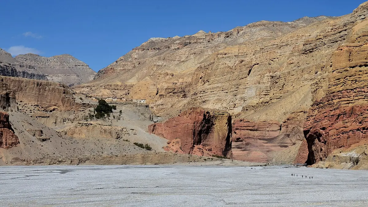 A wide, dry, pale stony riverbed under a blue sky, with bare cliffs of yellow, brown and red rock rising behind it, and a few tiny people walking across