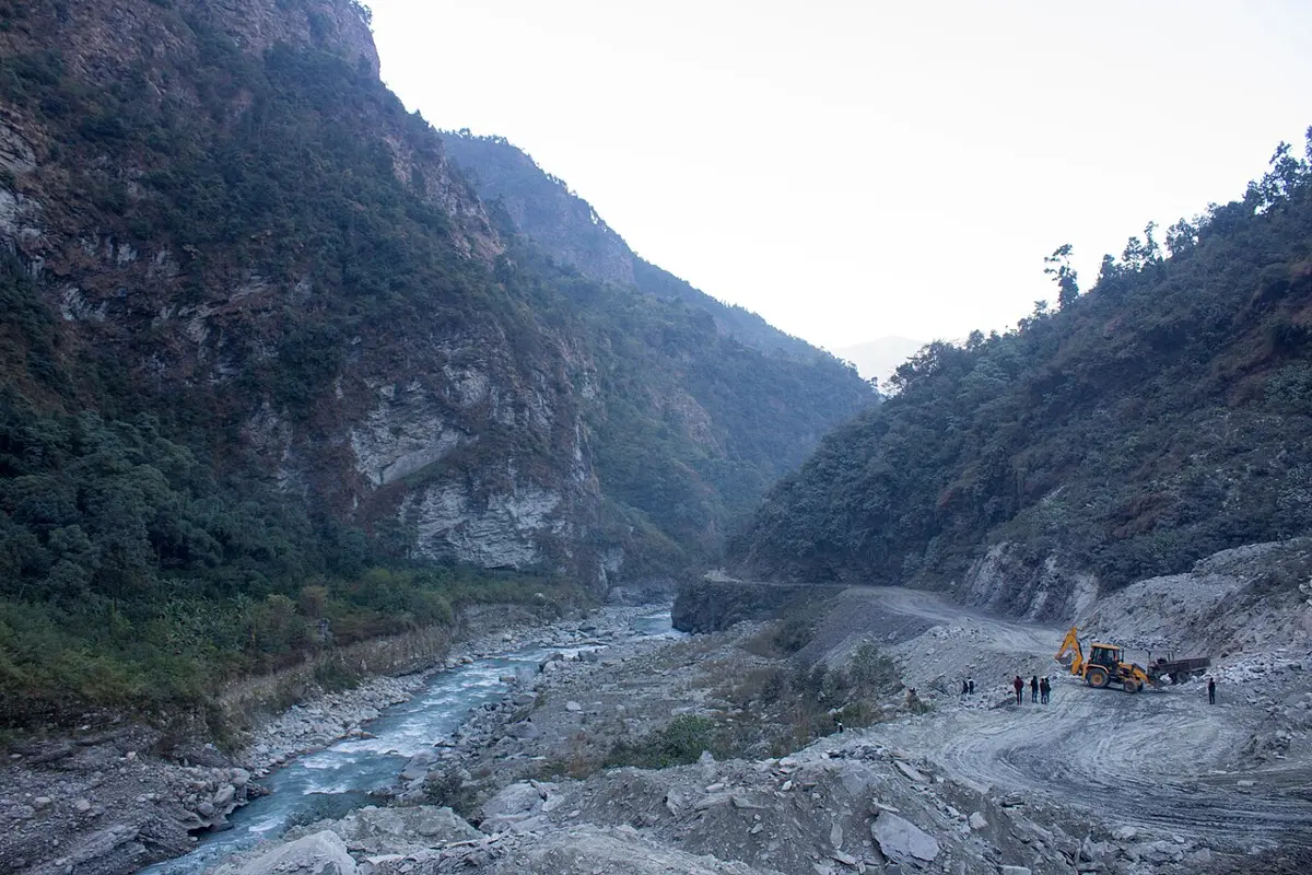 A narrow valley between steep, wooded mountainsides with gray cliffs, a blue-green river running over a bed of gray boulders, and a road being built on the right
