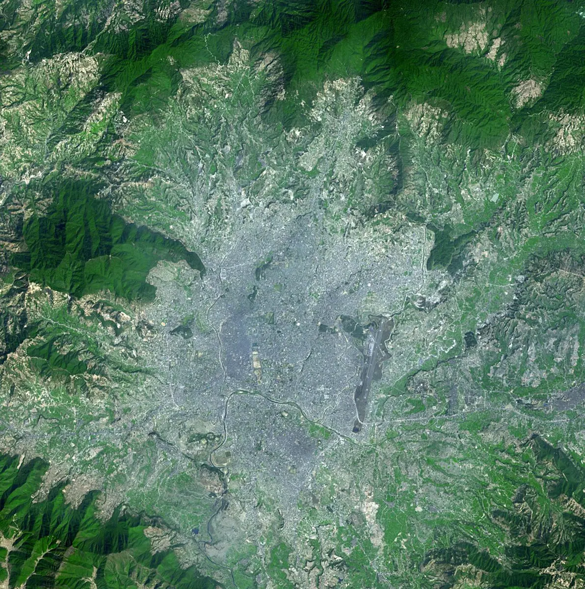 A satellite picture of a wide, flat valley ringed by green, forested hills; the middle of the valley is covered by a gray patch of city that spreads out along roads into the green
