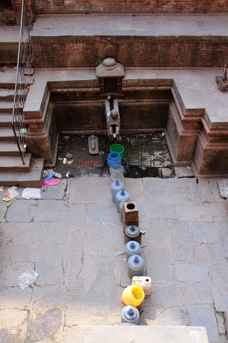 Looking down into a sunken stone spout built of brick and stone, with a thin stream of water falling from its carved spout; a long line of plastic jars and buckets stretches away from it across the paving, waiting to be filled