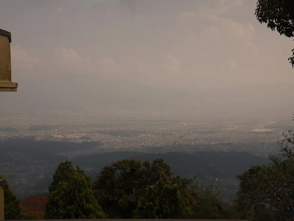 Looking down from a forested hillside over a wide valley filled with buildings, all under a thick brownish haze that hides the hills on the far side