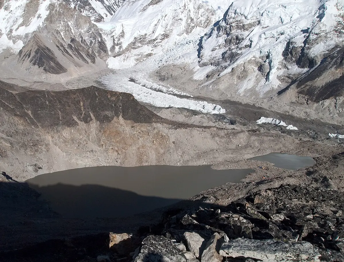 Snow-covered mountains above a jumbled white river of ice that pours down into a valley of gray rock and rubble, with a brown pond in the foreground