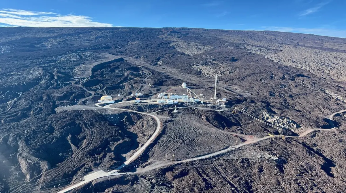 Seen from the air, a small cluster of white buildings and a tall mast in the middle of a huge slope of dark, bare lava, with a road winding up to them