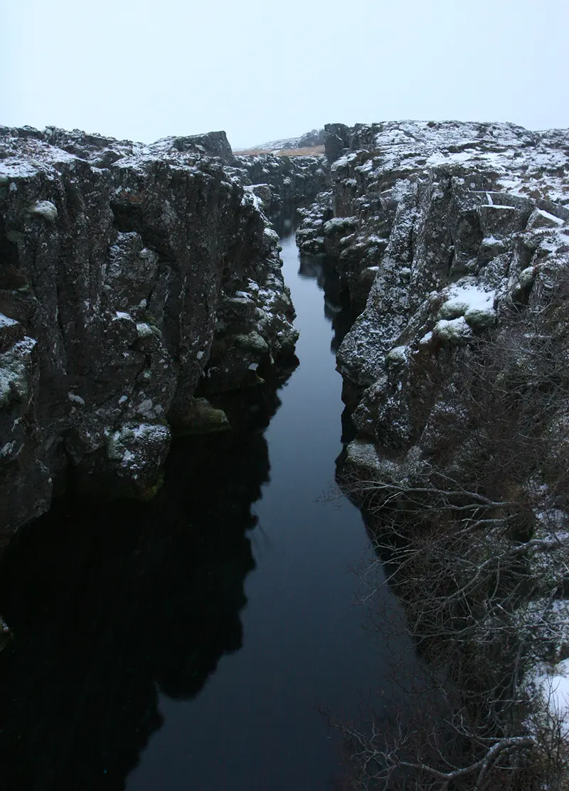 A narrow, straight crack between two walls of dark lava rock with snow on top, filled with still, dark water