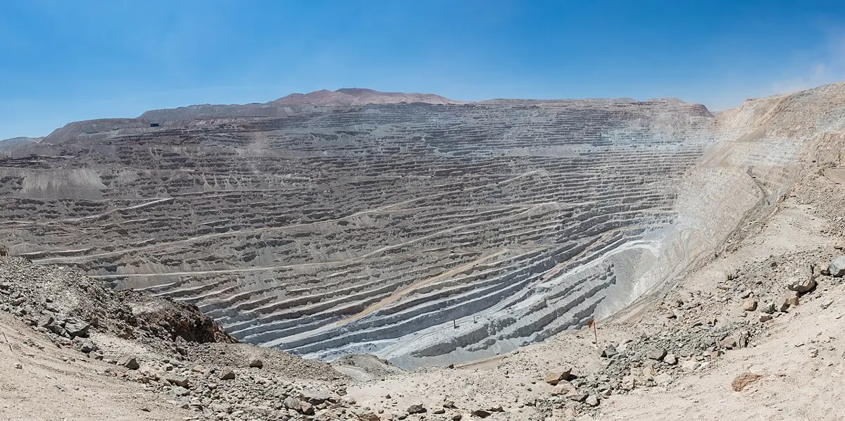 A gigantic pit in a desert, its gray walls cut into dozens of steps with tiny roads zigzagging down them, under a blue sky
