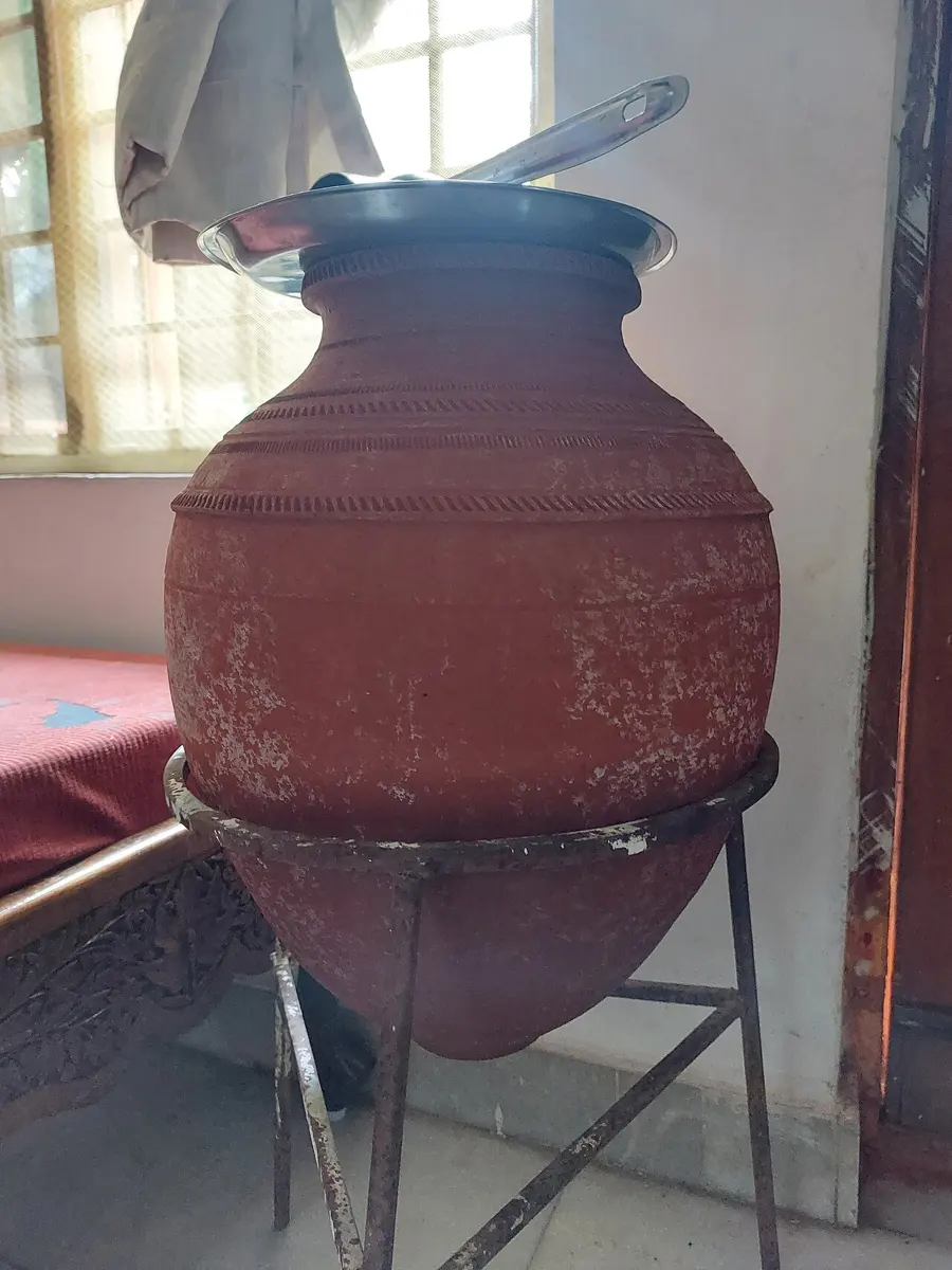 A round, reddish-brown clay water pot on an iron stand, covered with a steel plate, its outside patchy with white