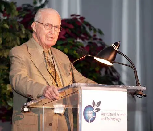 An old man with glasses, in a jacket and tie, speaking at a lectern