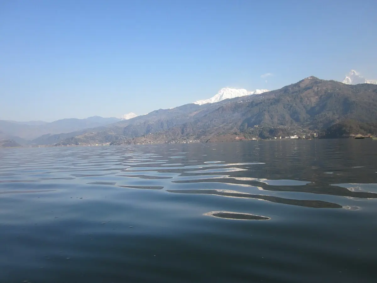A wide lake with small ripples, hills and a snowy peak behind; the water near the camera is dark, the far water reflects the hills and the pale sky