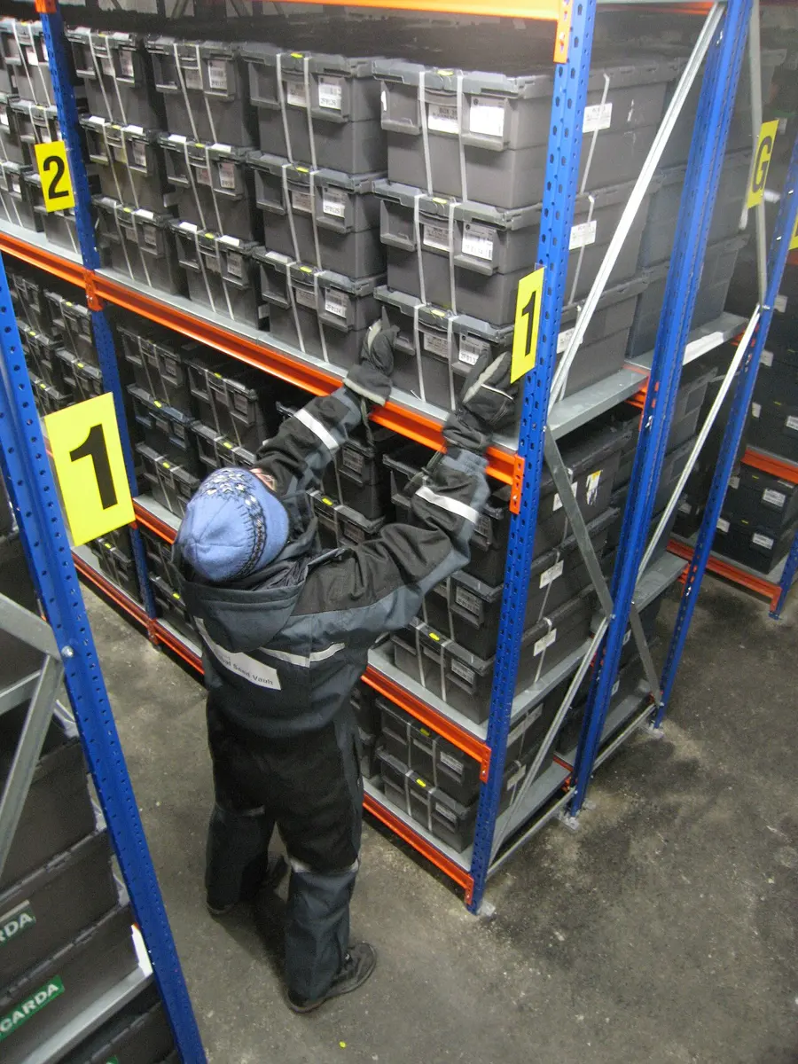 A person in a thick winter suit and hat reaches up to tall metal shelves stacked with sealed black boxes of seeds
