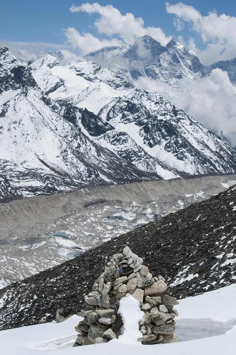 A wide glacier covered in gray stones, with patches of bare ice, between a long, steep-sided ridge of gray stones and snowy mountains