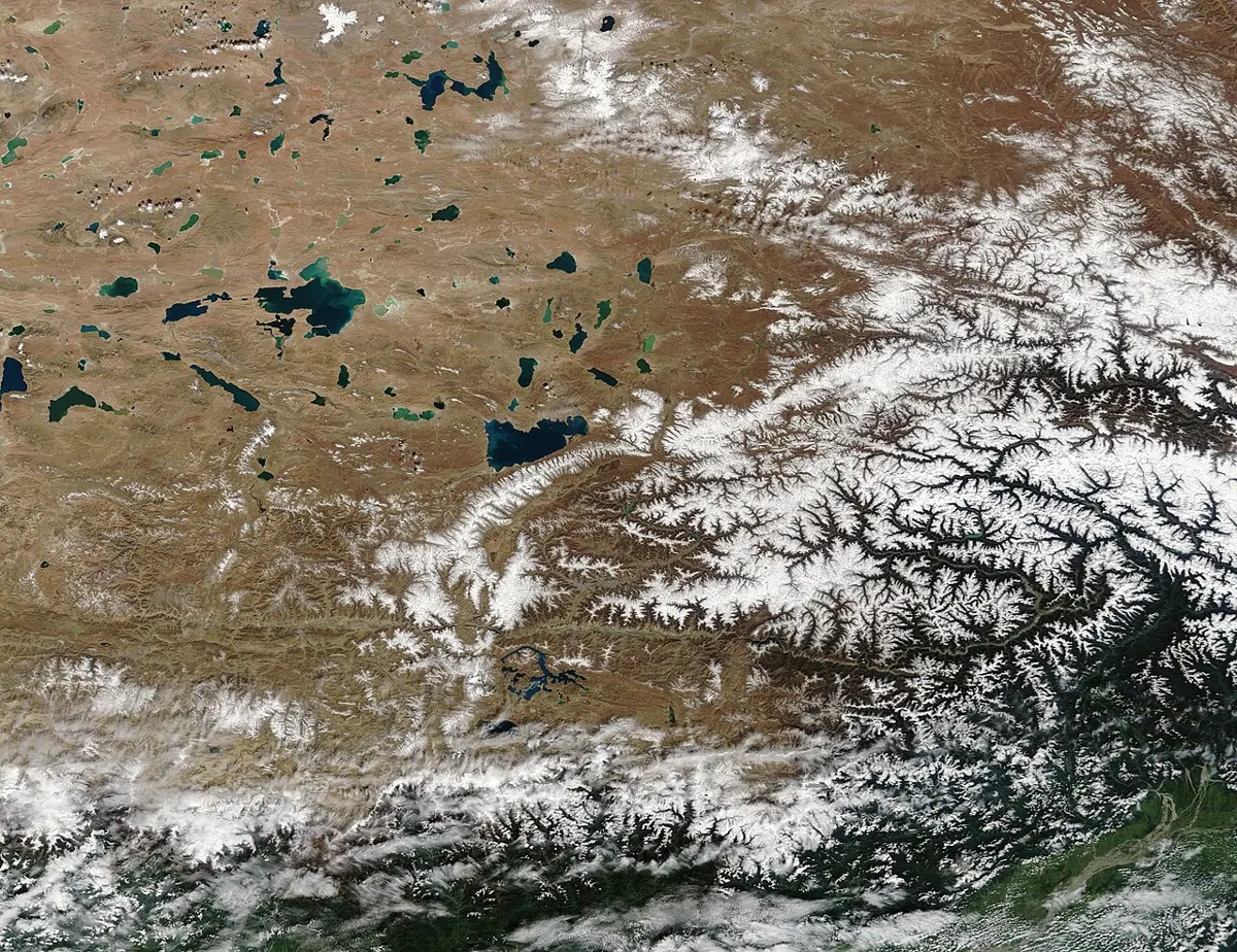 A satellite view of a high brown plain dotted with blue lakes in the upper left, crossed by a long band of snowy mountains; in the lower right, below the mountains, dark green forested valleys