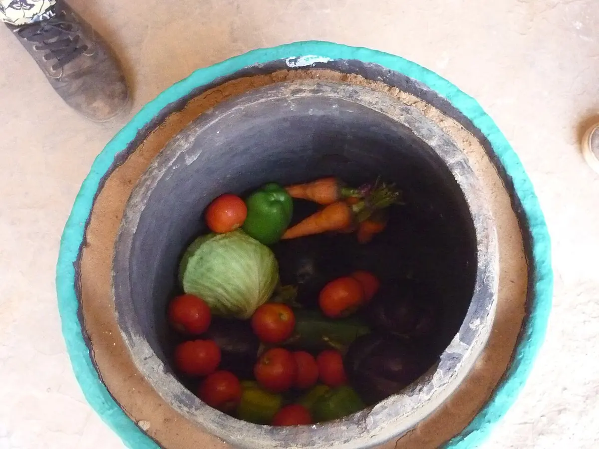 Looking down into a clay pot set inside a bigger one, with a ring of sand between them; inside are tomatoes, peppers, carrots and a cabbage