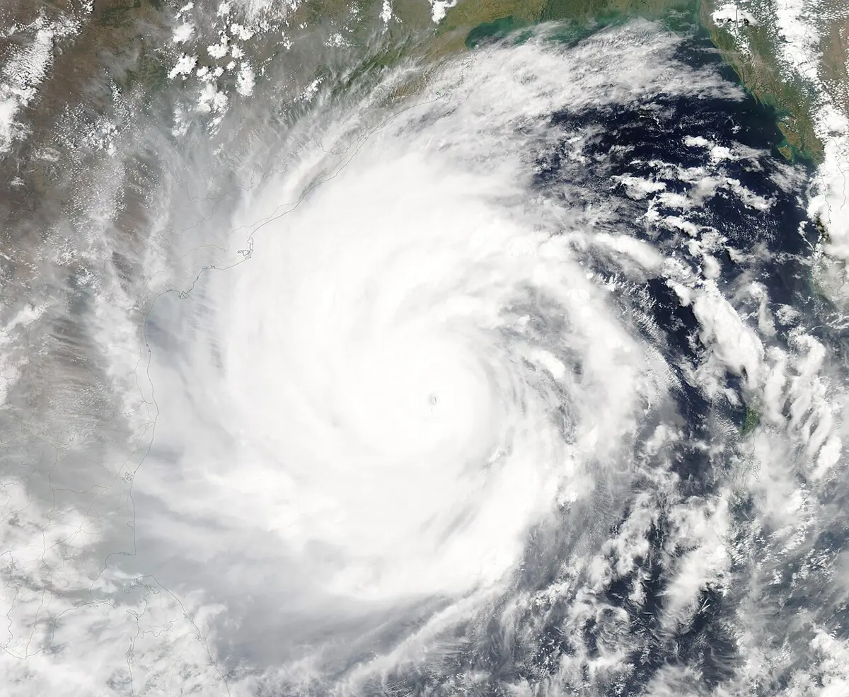 A huge swirl of white cloud, with a tiny dark eye at its center, covering most of a dark blue sea, with the brown and green coasts of land at the top and sides