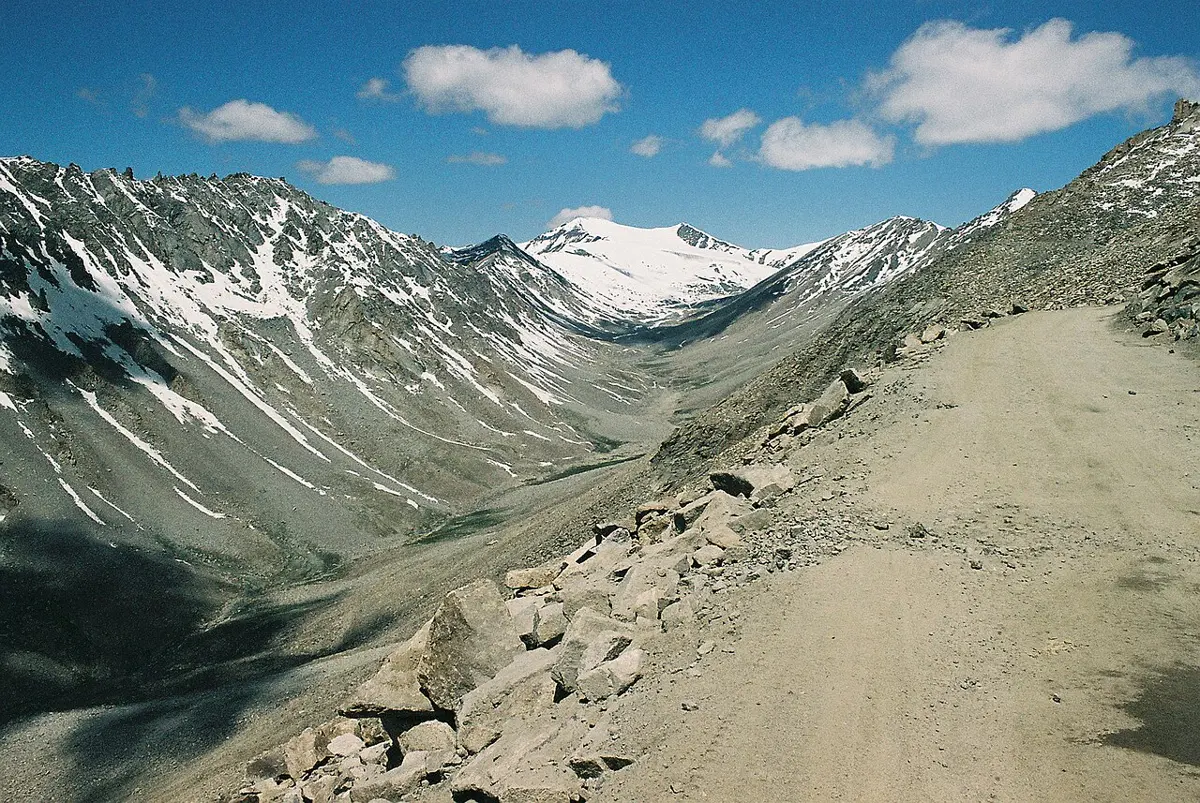 A wide valley between gray, snow-streaked mountains, with a broad, rounded floor of gravel, seen from a dirt road above it