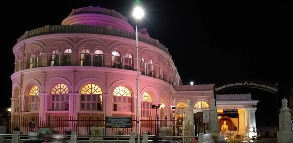 A large round building of two floors with rows of tall arched windows, lit pink and gold at night, behind a railing