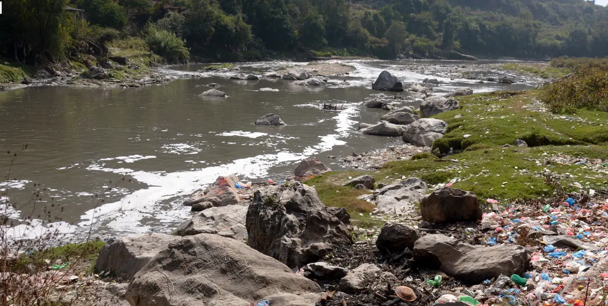 A shallow river flowing past large rocks; on the grassy bank in front, a wide spread of plastic bottles, bags and wrappers