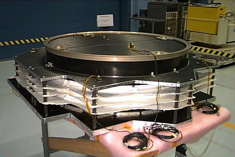 A round spacecraft part on a stand, with several thin sheets stacked one above another, separated by gaps and held apart by posts