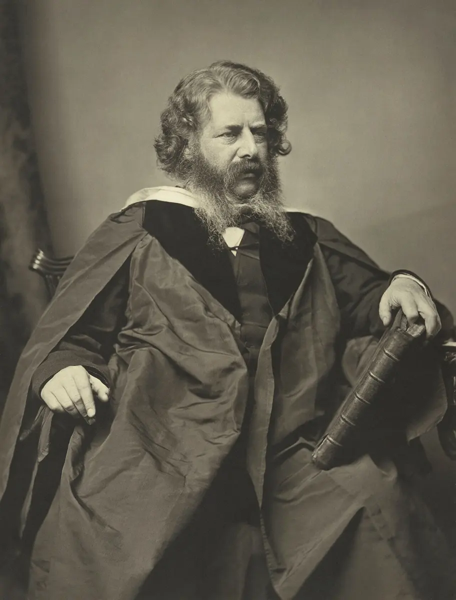 An old black-and-white photograph of a seated man with curly hair and a full beard, in a dark academic gown, one hand resting on a large book