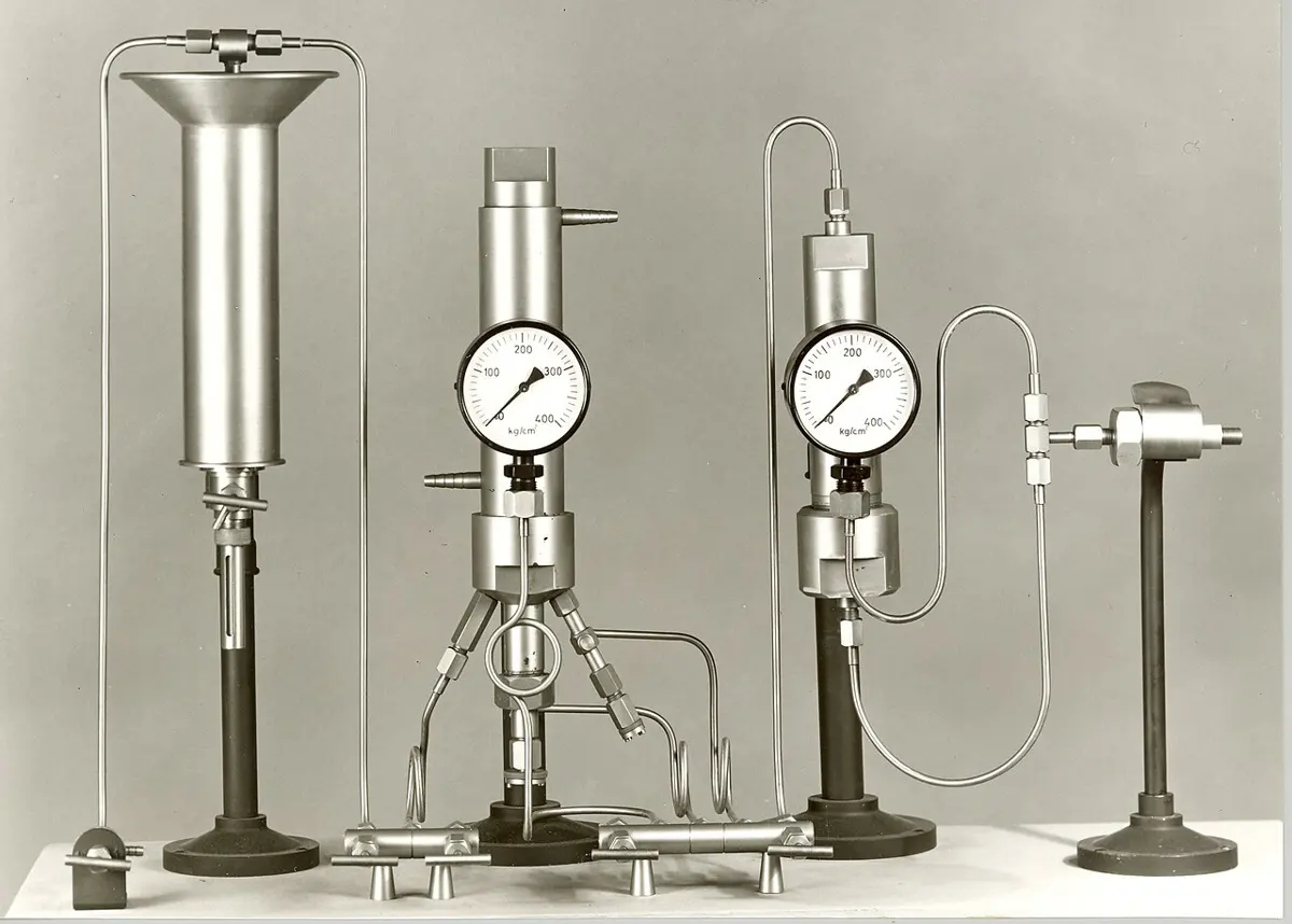 A black-and-white photograph of laboratory apparatus on a bench: tall steel cylinders on stands, two round pressure gauges, and thin metal pipes joining them