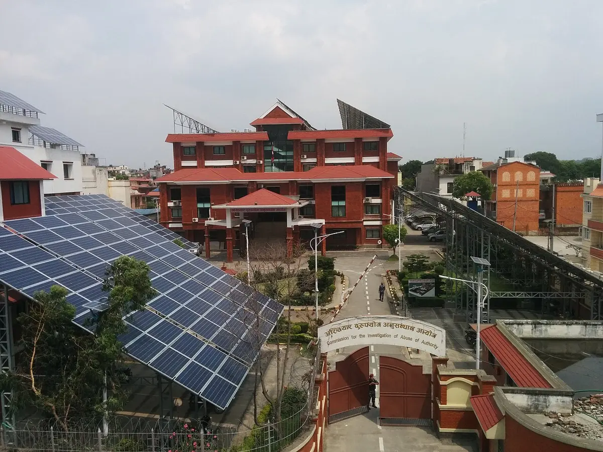 Rows of solar panels on a tall metal frame shading a garden and car park in front of a red-brick government building in Kathmandu, with more panels on the building’s roof
