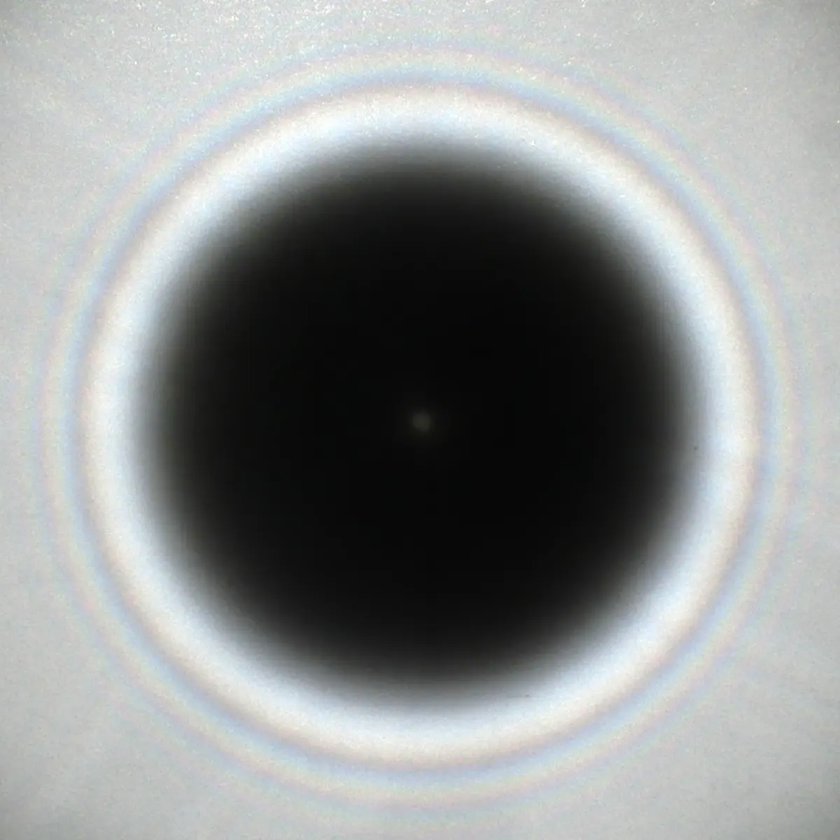 A round dark shadow on white paper, ringed by faint colored circles, with a small faint bright dot exactly in its center