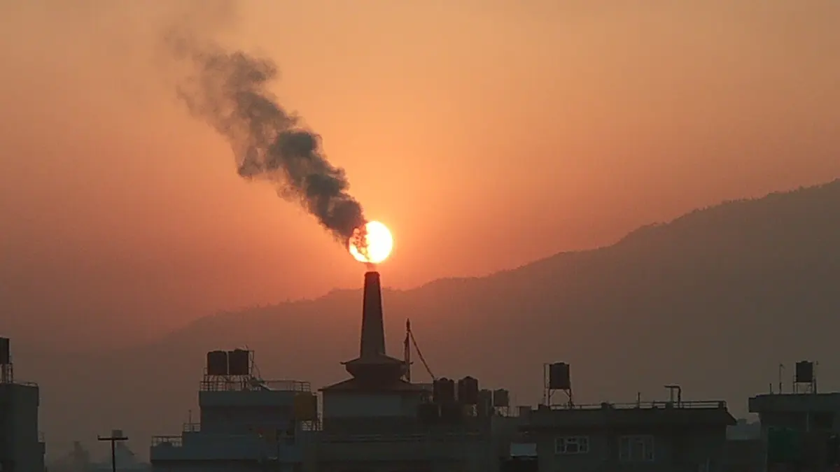 A brick kiln chimney silhouetted against an orange morning sky, with a plume of dark smoke streaming from its top across the rising Sun
