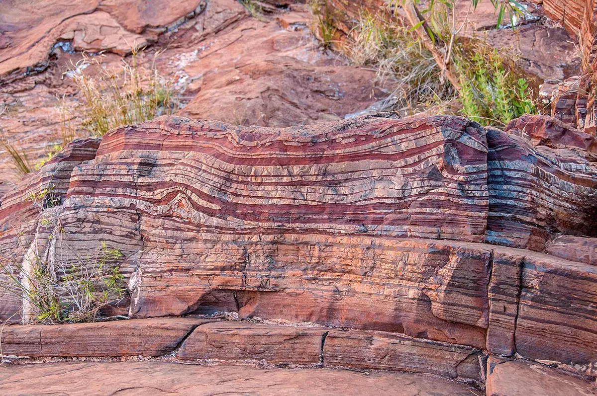 A cliff of rock in thin, wavy stripes of dark red, purple-gray and orange