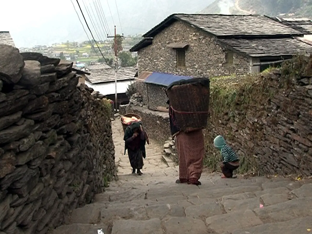 A stone-paved lane between houses with walls of rough stone and roofs of gray stone slates; people carry big baskets along the lane