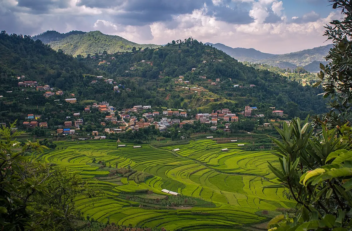 A village of red and white houses on a green hillside, with terraced rice fields in curving steps below it, and forested hills behind under a cloudy sky