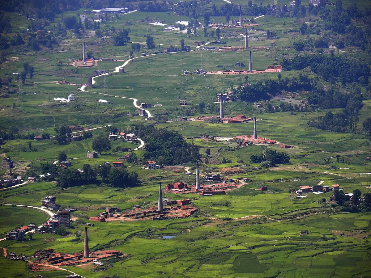 Green rice fields seen from a hillside, dotted with about a dozen tall, thin brick chimneys, each beside a patch of red brick kiln and stacked bricks