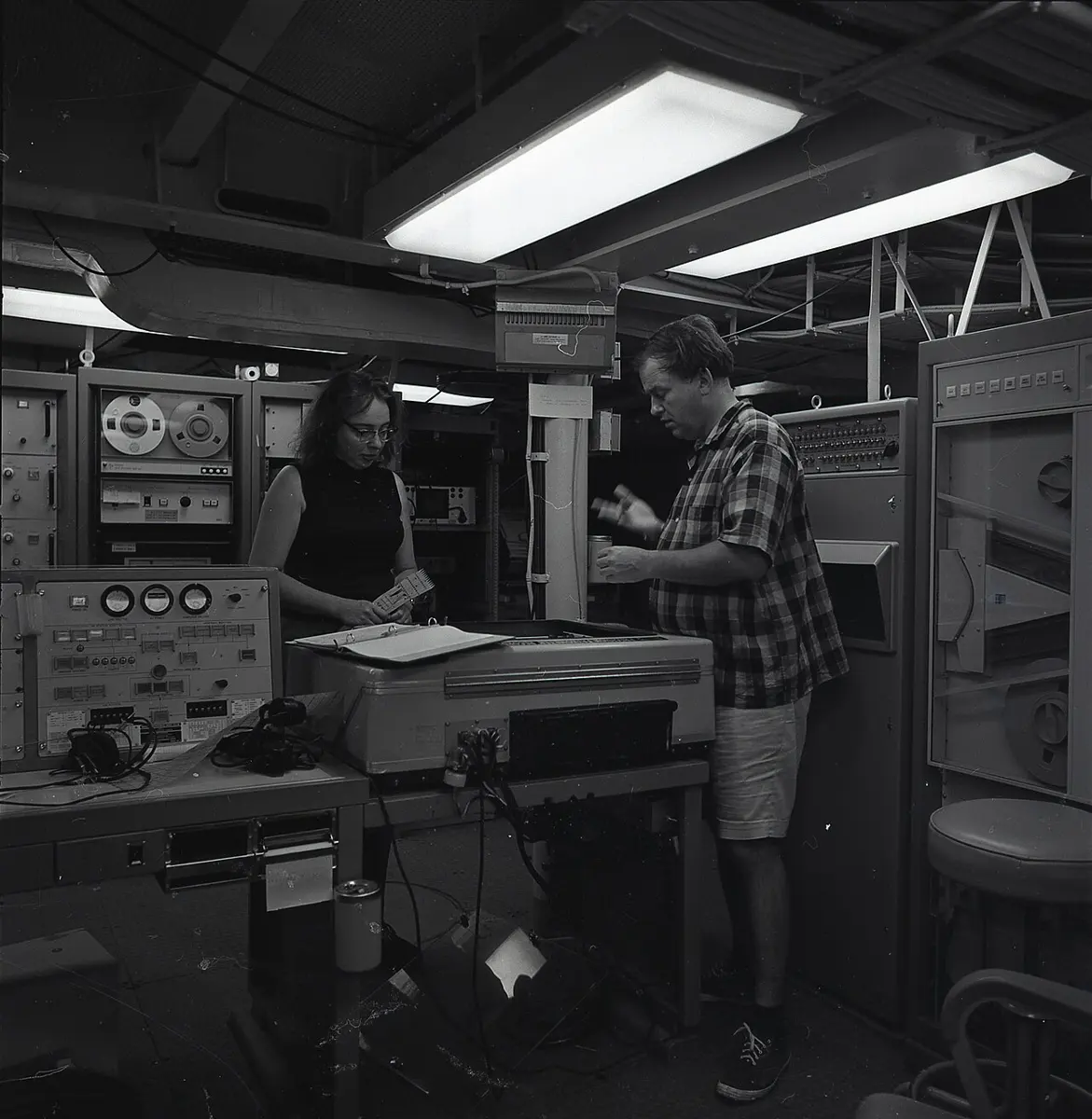 A black-and-white photograph of a man and a woman standing among racks of electronic equipment on a ship, looking at papers