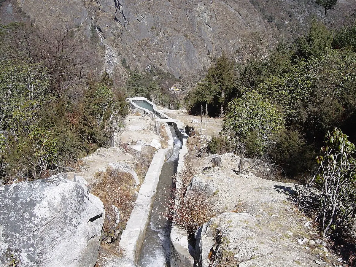 A narrow concrete-lined canal of rushing water running along a rocky hillside to a small tank, with a steep mountainside behind