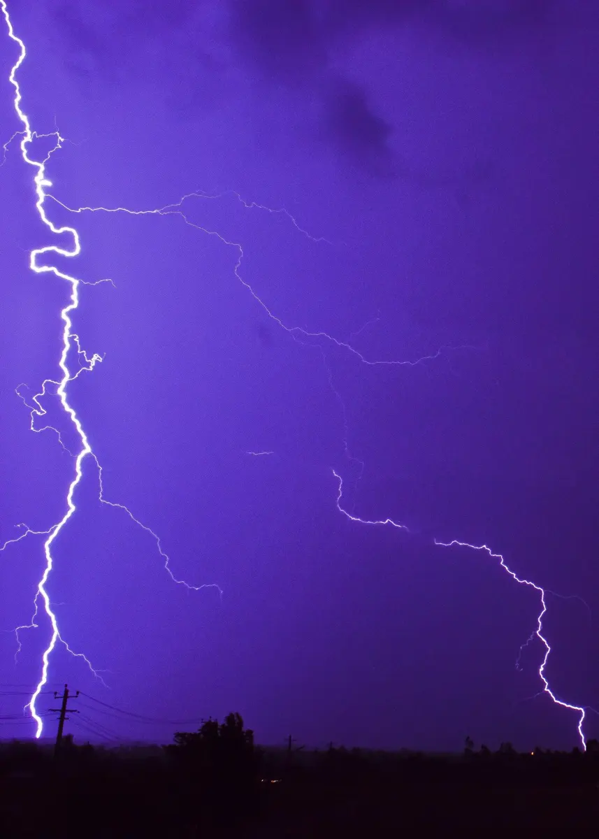 Bright, branching bolts of lightning from a purple night sky down to a dark, flat landscape with an electricity pole