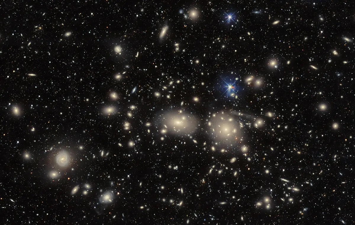 Hundreds of fuzzy oval and spiral galaxies scattered across a dark sky, with two large bright oval galaxies near the middle