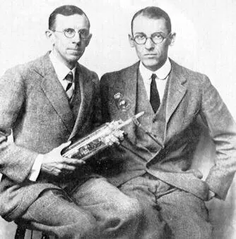 An old black-and-white photograph of two men in suits and round glasses, seated side by side; the one on the left holds a glass tube with metal parts inside
