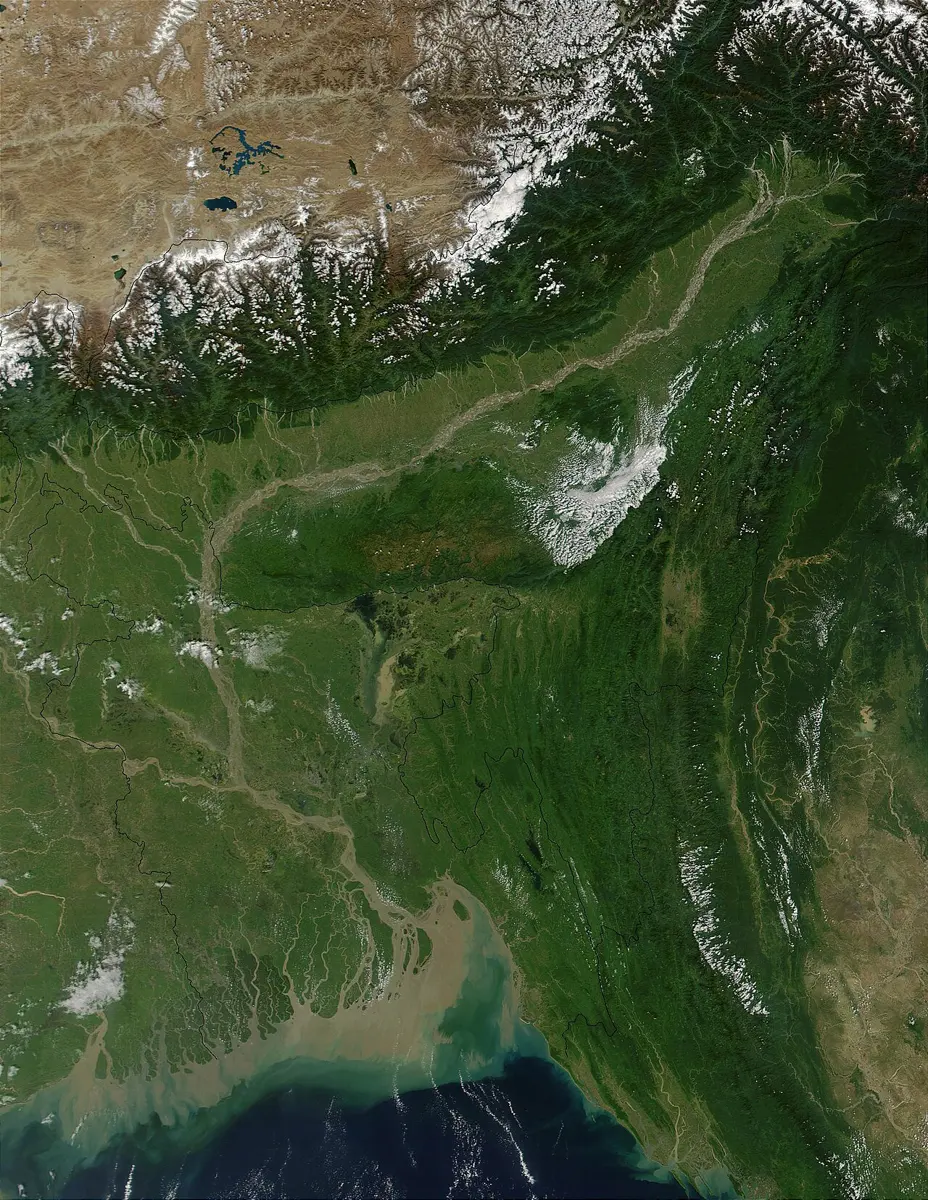 A satellite view of snowy Himalayan peaks and the brown Tibetan Plateau at the top, green plains in the middle crossed by pale rivers, and at the bottom a delta where brown, muddy water spreads into a dark blue sea