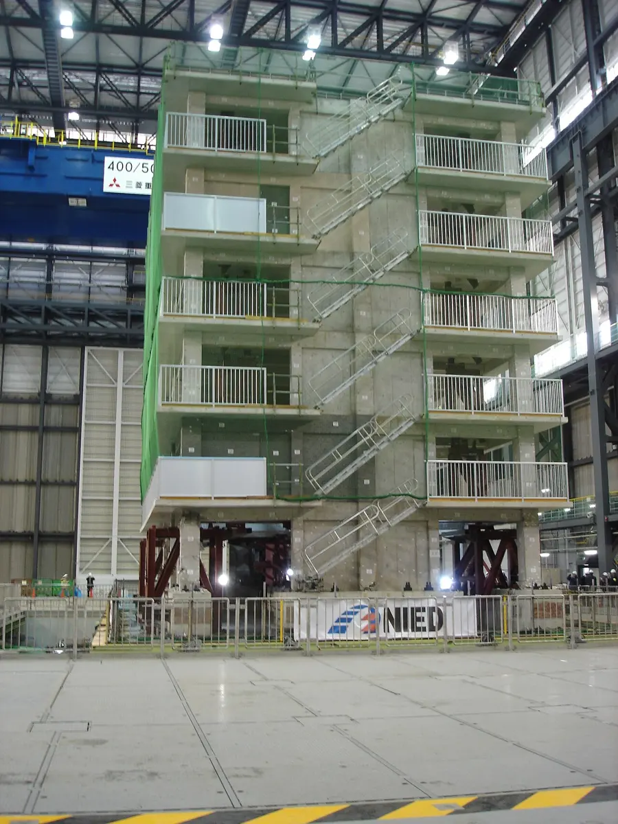 A full-size six-storey concrete building with balconies and outside stairs, standing inside a huge hall on the flat floor of the E-Defense shake table