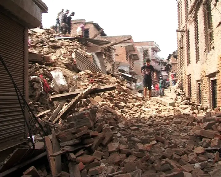 A street in Bhaktapur filled with a heap of fallen bricks and broken timber, with people standing on the rubble and houses still standing on either side